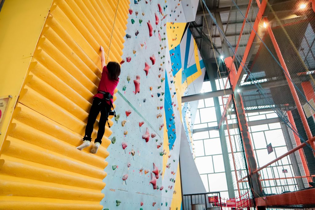 A girl in a harness climbs up a bright yellow indoor climbing wall with lots of rock walls and other activities in the background. The TREK Fuller Foundation Scholarship pays for two youth to participate in adventure-based programming, such as indoor rock climbing. 