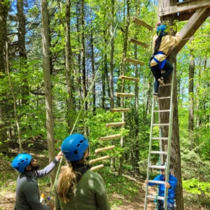 A young woman in a harness and helmet climbs a ladder leading against a tree as part of a challenge course while a LUK staff wearing a helmet is on belay, another staff member secures the ladder, and another staff supervises