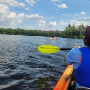 A LUK staff kayaking on a lake with a blue sky and trees in the background. Another kayak is also visible in the near distance.