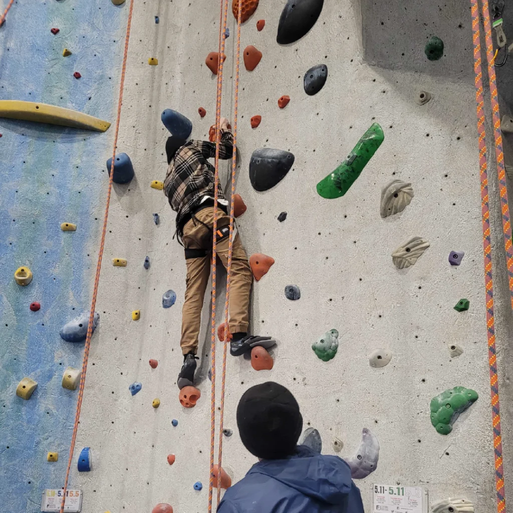 A young adult climbs an indoor climbing wall with a LUK staff on belay