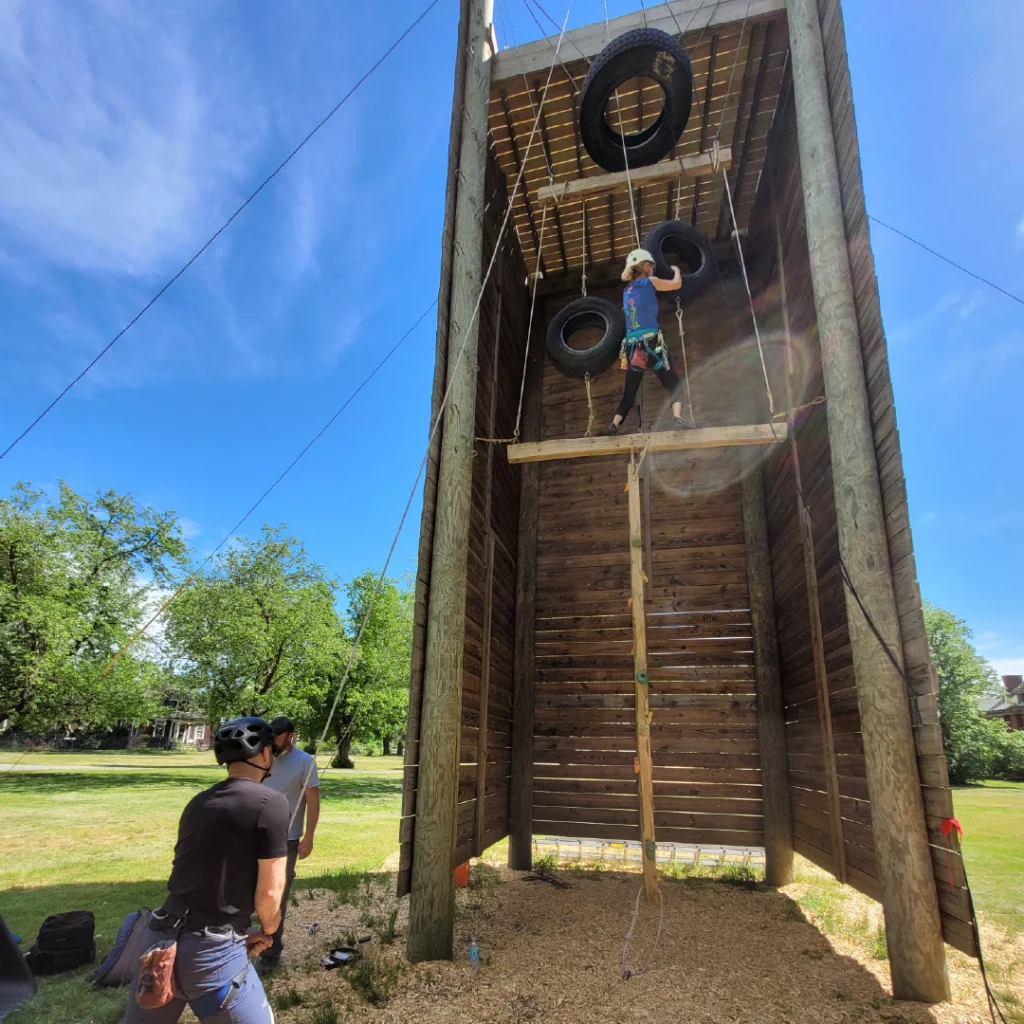A woman balances on a challenge course holding onto a tire swing, with a LUK staff on belay