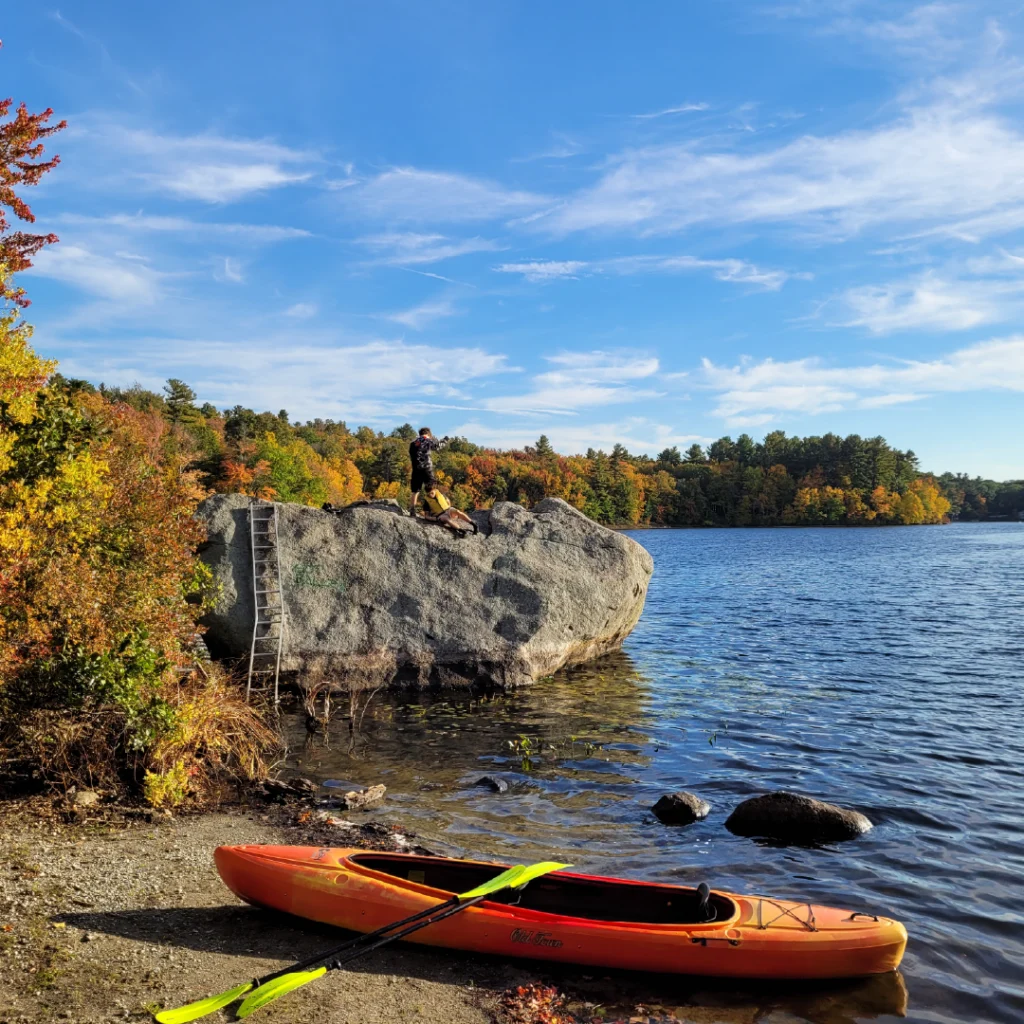 A kayak and paddles rests on the shore of a lake next to autumn foliage as youth explore the natural surroundings