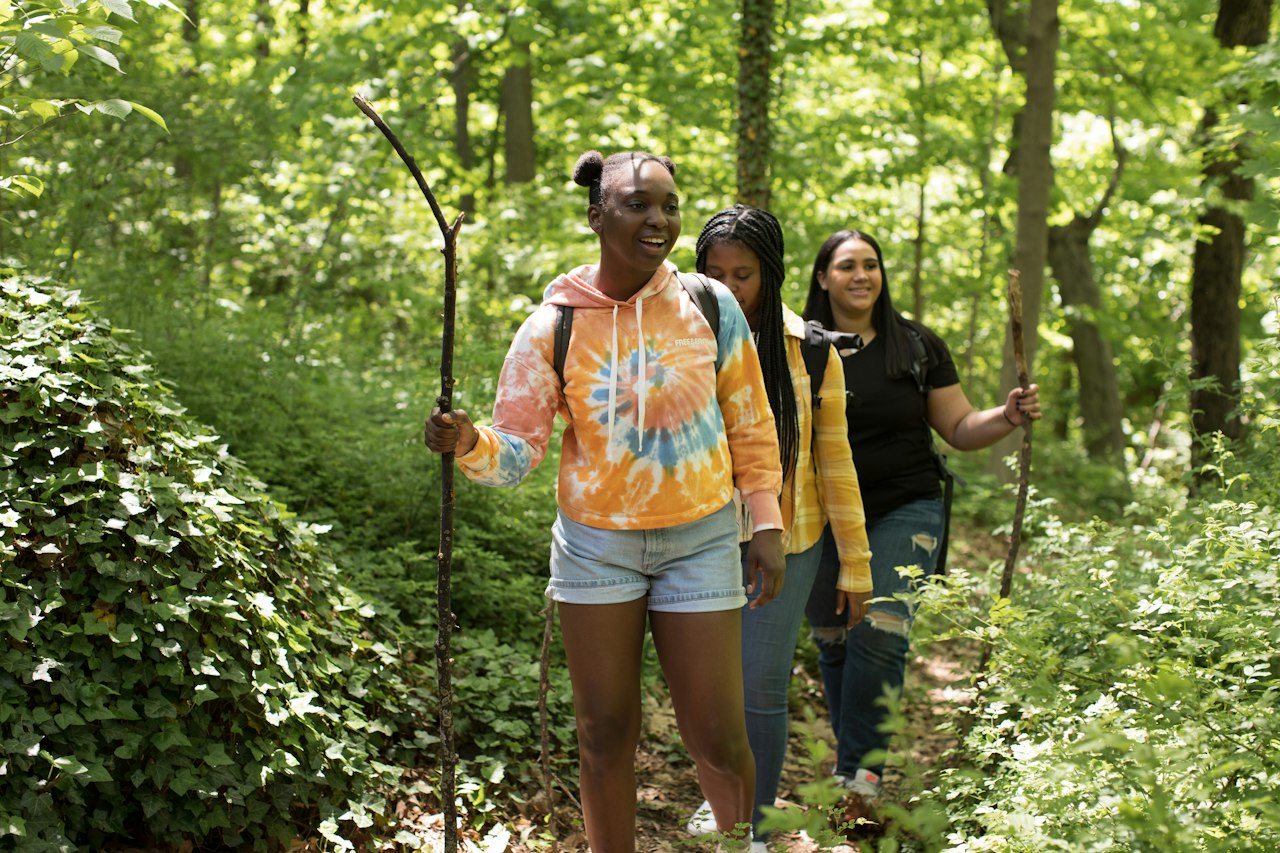 Three high school aged girls walk through a path in the woods. Hiking is a part of LUK Adventure-Based Program services.